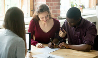 Photo of three people sitting around a table working, one with a laptop, and two looking at the same paper.